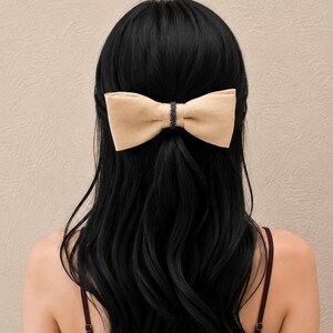 May include: A beige bow hair clip with a black beaded centre secures a section of long, dark, wavy hair at the back of the head. The background is a plain, neutral colour.