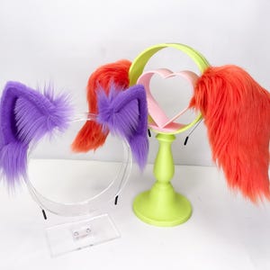 May include: A display of quirky headwear, including purple cat ears and orange faux fur accents. A lime green stand holds a pink heart-shaped frame with orange fur. The items are set against a white backdrop.