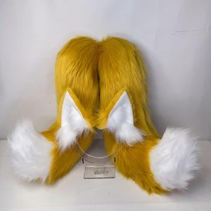 Könnte beinhalten: Ein Set aus Fuchsohren und -schwanz, hergestellt aus gelb-orangefarbenem und weißem Kunstfell. Die Ohren haben weiße Innenflächen, und der Schwanz hat eine flauschige weiße Spitze. Für Cosplay oder Kostümzwecke.