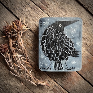 May include: A rectangular wooden plaque featuring a painted black crow on a grey background. The crow has white outlined feathers, a single white eye, and stands on thin legs. The background includes white star shapes and three small circles.
