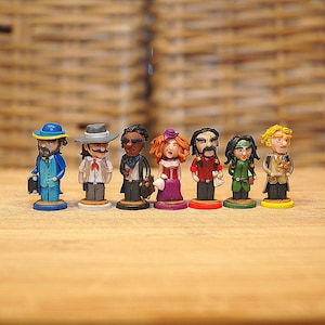 Op de afbeelding: Een verzameling van zeven kleine, handgeschilderde beeldjes die verschillende personages uitbeelden. De beeldjes dragen kleurrijke outfits, waaronder hoeden, vesten en jurken. Ze staan op kleine, ronde voetstukken en zijn gerangschikt op een houten oppervlak.