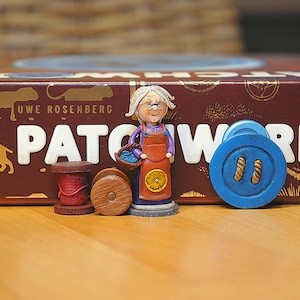 Könnte beinhalten: Ein Brettspiel namens "Patchwork" von Uwe Rosenberg. Das Spiel hat eine farbenfrohe Schachtel mit einer Frau, die einen Korb mit Knöpfen hält. Es gibt auch Garnrollen und einen großen blauen Knopf.
