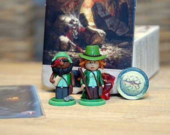 May include: Two green and brown figurines of people wearing green hats and holding a staff and a book. The figurines are standing on green bases. A green and brown compass token is in the background.
