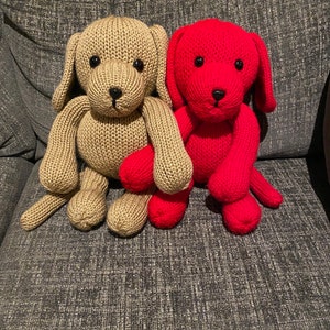 May include: Two knitted dog toys, one beige and one red, sitting on a grey sofa.