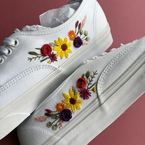 May include: White canvas sneakers with colorful floral embroidery. The embroidered flowers include yellow sunflowers, red roses, and purple accents. The shoes have white laces and a white rubber sole.