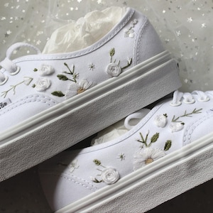 May include: White canvas sneakers with floral embroidery. The shoes feature embroidered white flowers, green leaves, and delicate stems. The sneakers have white laces and a white rubber sole. The brand name "Vans" is visible on the side of the shoe.