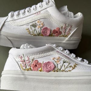 May include: White canvas low-top sneakers with pink and green embroidered flowers. The shoes have a white rubber sole and a white lace-up closure.