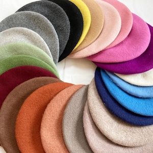 May include: A collection of circular felt berets arranged in a fan-like pattern. The hats feature a wide spectrum of colours including shades of grey, black, yellow, beige, pink, magenta, purple, white, blue, navy, brown, orange, and green.