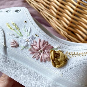 May include: White canvas shoe with embroidered floral details. The embroidery features pink, yellow, and white flowers, along with green leaves and small bees. The shoe has a white rubber sole and eyelets.
