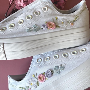 May include: White canvas sneakers with floral embroidery in pastel colors. The embroidered flowers include pink roses, purple buds, and white daisies. The sneakers have white soles and eyelets.