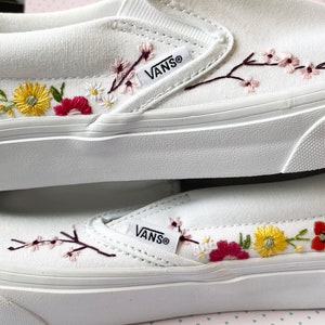 May include: White slip-on sneakers with embroidered floral designs. The sneakers feature colorful flowers in red, yellow, and pink, along with green leaves and brown stems. The brand name "VANS" is visible on the side.