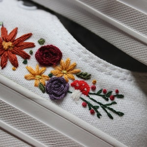 May include: White canvas shoes with embroidered flowers in shades of red, orange, yellow, purple, and green. The flowers are arranged in a row along the side of the shoe.