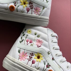 May include: White high-top sneakers with colorful floral embroidery. The embroidered flowers include yellow, pink, and orange blooms with green leaves. The sneakers have white laces and a white sole.