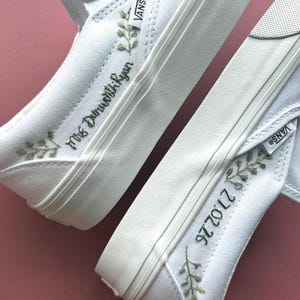 May include: White Vans slip-on sneakers with green embroidered floral details. One shoe features "Mrs Dunworth-Ryan" in green script, the other displays "27.02.26". The shoes have white soles and a canvas upper, perfect for casual wear.