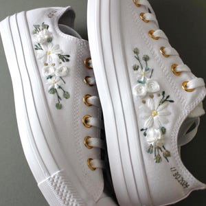 May include: White canvas sneakers with gold eyelets and embroidered white flowers and green leaves. The right shoe has the date "11.30.2024" embroidered on the side.