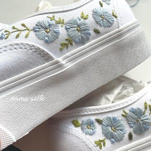 May include: White canvas sneakers with embroidered light blue flowers and green leaves. The floral design is on the outer side of the shoe. The sneakers have a thick white sole and are sitting on white tissue paper.