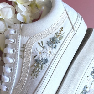 May include: White canvas sneakers with floral embroidery in shades of blue, green, and white. The sneakers feature a classic lace-up design and a white rubber sole. The initials "A&W" are embroidered on the side.
