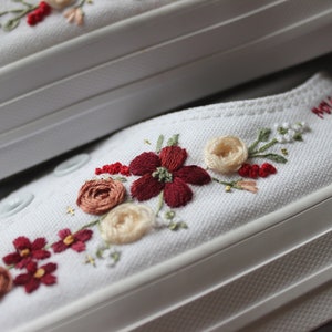 May include: White canvas shoes with embroidered flowers in shades of red, pink, and cream. The flowers are arranged in a line along the side of the shoe.