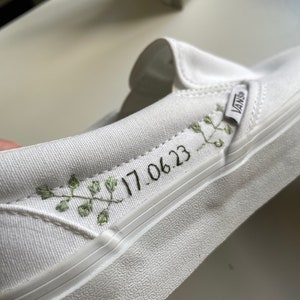 May include: White slip-on shoe with the brand name "Vans" on the side. The shoe features embroidered green leaves and the date "17.06.23" in black. The shoe has a white sole and a canvas upper.