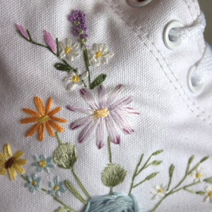 May include: White canvas shoes with embroidered flowers in shades of purple, pink, yellow, blue, and green. The flowers include daisies, lavender, and a blue rose.