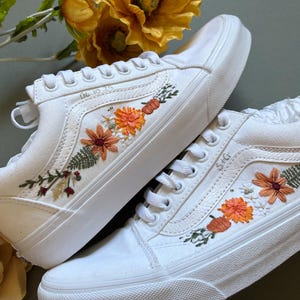May include: A pair of white canvas sneakers with orange, red, and green embroidered flowers. The sneakers have a classic low-top design and are decorated with a floral pattern.
