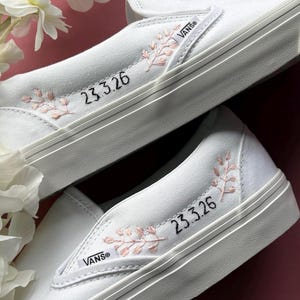 May include: White slip-on sneakers with embroidered pink floral accents and the date "23.3.26" in black. The sneakers feature the Vans logo on the side and a white sole.