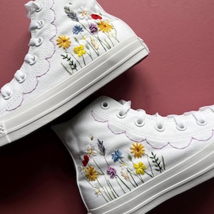 May include: White high-top sneakers with embroidered floral designs in yellow, red, purple, and blue. The sneakers have a scalloped edge and white laces. The background is a solid maroon color.