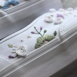 May include: Close-up of white canvas sneakers with embroidered floral designs. The embroidery features various colors, including green, purple, and white, creating a delicate and artistic look. The sneakers have a thick, textured white sole.