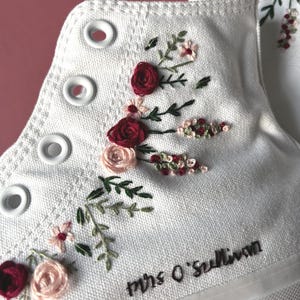 May include: White high-top canvas sneakers with embroidered floral details. The embroidery features red and pink roses, small pink flowers, and green leaves. The text "Mrs O'Sullivan" is embroidered on the side.