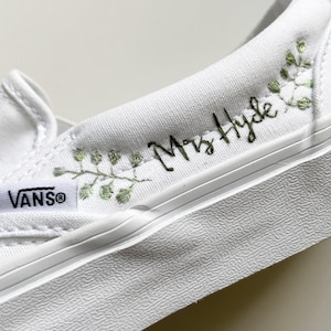 May include: White Vans slip-on sneakers with the Vans logo on the side. The shoe is customized with the text "Mrs. Hyde" in black script and embroidered green floral accents.