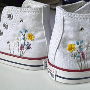 May include: A pair of white high-top sneakers with embroidered floral and bee designs. The sneakers have a red stripe along the sole and silver eyelets. The Converse All Star logo is visible on the heel.