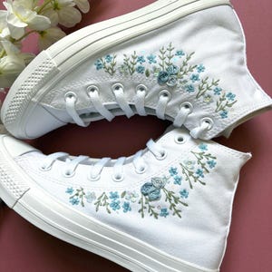 May include: White high-top sneakers with embroidered floral designs in shades of blue and green. The sneakers feature a classic lace-up design and a white rubber sole. The floral embroidery adds a delicate, handcrafted touch to the shoes.