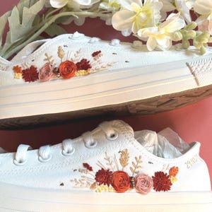 May include: White canvas sneakers with embroidered floral designs in shades of red, orange, and pink. The sneakers have white laces and soles. The date "29.10.21" is embroidered on one shoe.