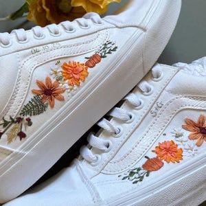 May include: White canvas trainers featuring embroidered floral patterns in orange, red, and green. The trainers have white laces and soles. The text "04.10.75" and "S+G" are embroidered on the sides. A unique and stylish footwear option.