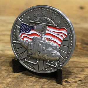 Puede incluir: Una moneda de desafío plateada que presenta un helicóptero con una bandera estadounidense en el fondo. La moneda tiene el texto "UH-1 HULSEY" y "61" en ella.