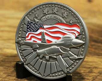 May include: A silver challenge coin featuring a raised image of an F-16 fighter jet flying over an American flag. The coin has a black stand and the text "F-16 Fighting Falcon" is inscribed on the coin.