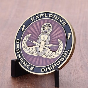 Könnte beinhalten: Eine runde, goldfarbene Münze mit dem Schriftzug "EXPLOSIVE ORDNANCE DISPOSAL" in schwarzer Schrift. Die Münze zeigt ein zentrales Emblem mit einem Stern, einer Bombe und Blitzen, vor einem violett-goldenen radialen Design. Die Münze wird auf einem schwarzen Ständer präsentiert.
