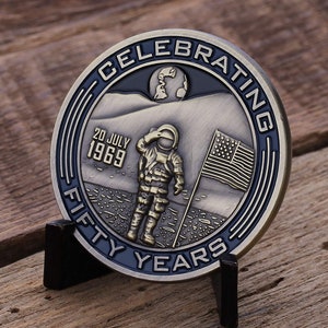 May include: A commemorative coin celebrating fifty years since the 1969 moon landing. The coin features an astronaut saluting the American flag on the moon, with the Earth in the background. The text reads "Celebrating Fifty Years" and "20 July 1969".