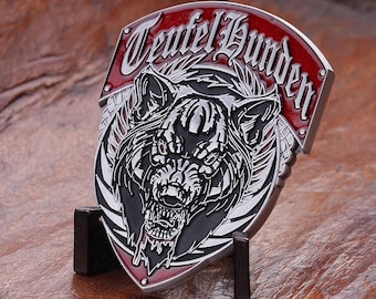 May include: A silver-colored metal coin with a detailed wolf head design. The coin has the words "Teufel Hunden" in a gothic font on a red banner. The coin is displayed on a small black stand.