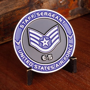 May include: A round coin featuring the words "Staff Sergeant" and "United States Air Force" in blue and white. The coin has a gray center with a star and stripes emblem, and the designation "E-5".
