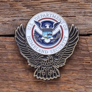 May include: A metal badge featuring the U.S. Department of Homeland Security seal. The circular emblem has a white border with the text "U.S. DEPARTMENT OF HOMELAND SECURITY". The seal includes a blue eagle and a shield, with a red and blue color scheme.