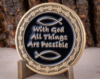 Puede incluir: Una moneda de color dorado con fondo negro y la inscripción "With God All Things Are Possible" rodeada de un diseño de alambre de púas. Dos símbolos de peces estilizados se encuentran a cada lado del texto. La moneda está en un soporte dorado.