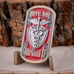 May include: A rectangular metal challenge coin with the words "DEVIL DOC" and "CORPSMAN UP" in silver lettering. The coin features a red cross, a silver caduceus symbol, and a gold rope border. The coin is displayed on a small stand.