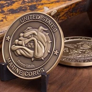 May include: A gold-colored challenge coin with a bulldog head in the center. The coin reads "United States Marine Corps" around the edge and "Semper Fidelis" below the bulldog.
