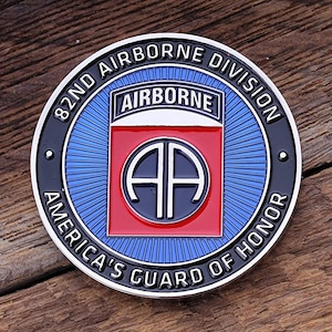 Puede incluir: Una moneda metálica redonda con un diseño azul, rojo y negro. La moneda presenta el texto "82ND AIRBORNE DIVISION AMERICA'S GUARD OF HONOR" y la palabra "AIRBORNE". El centro tiene un cuadrado rojo con las letras "AA".