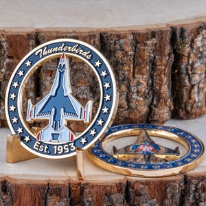 May include: Two gold-colored commemorative coins featuring a blue and white jet aircraft design. The coins have a circular shape with a blue border, white stars, and the text "Thunderbirds Est. 1993". One coin is displayed upright on a gold stand.