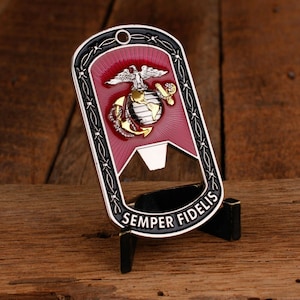 Puede incluir: Un abridor de botellas de metal con un emblema rojo y dorado que presenta un águila, un globo terráqueo y un ancla. El abridor tiene las palabras "Semper Fidelis" y se muestra en un pequeño soporte negro.