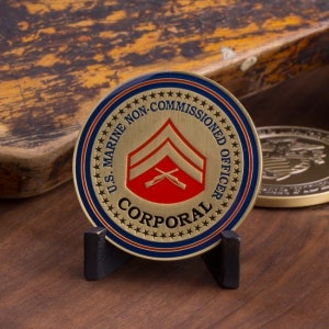 May include: A gold-colored coin with a blue and red design. The coin features the text "U.S. MARINE NON-COMMISSIONED OFFICER" and "CORPORAL" in a circle around a red and gold chevron with crossed rifles. The coin is on a black stand.