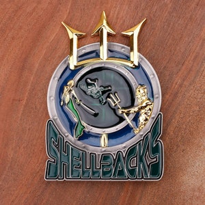 May include: A detailed, colorful metal pin featuring a trident, a mermaid, and a figure with a trident. The pin has a blue and silver background with the word "SHELLBACKS" in green and white lettering. The pin is on a wooden surface.