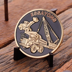 May include: A round, bronze-colored coin with a black center, featuring construction vehicles and the word "ESSAYONS" in red. The coin is displayed on a small, dark stand against a wooden background.
